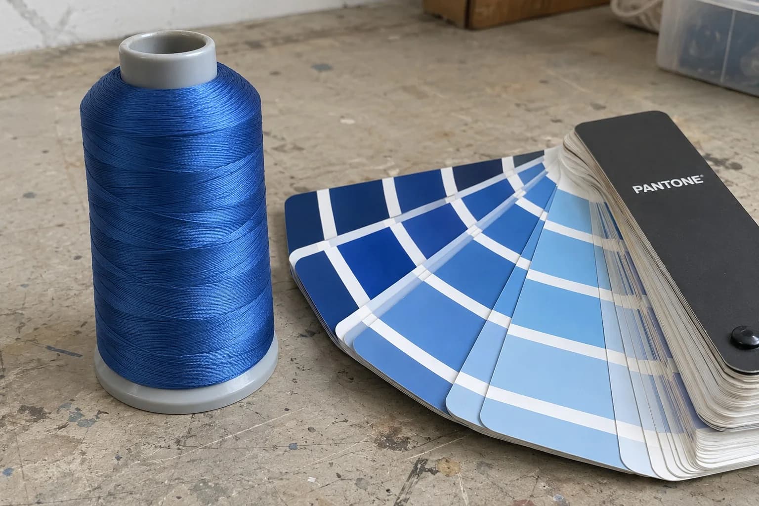 A cone of embroidery thread beside an open Pantone Coated fan deck, matching a blue thread to its nearest Pantone chip